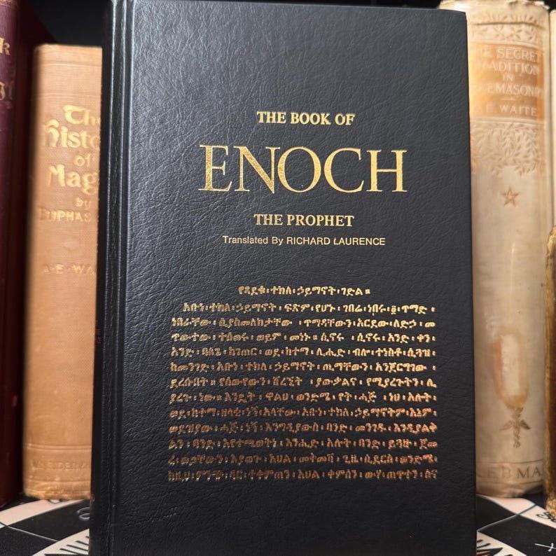 Puede incluir: Un libro de tapa dura negro titulado "The Book of Enoch" con letras doradas. El texto incluye "The Prophet" y "Translated By Richard Laurence." Otros libros son visibles en el fondo.