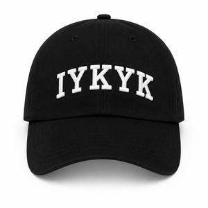 Puede incluir: Una gorra de béisbol negra con el texto IYKYK bordado en letras mayúsculas blancas en el panel frontal. La gorra tiene una visera curva, una corona estructurada con un botón superior y ojales cosidos para ventilación en los paneles superiores.