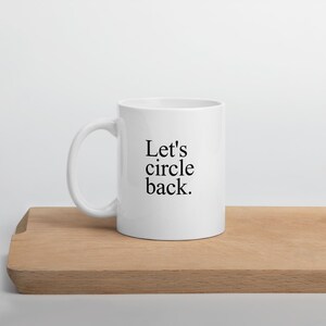May include: A white ceramic mug with a handle on the left side. The text "Let's circle back." is printed in black, serif font on the front of the mug. The mug has a smooth, glossy finish and a cylindrical shape with a flat base.