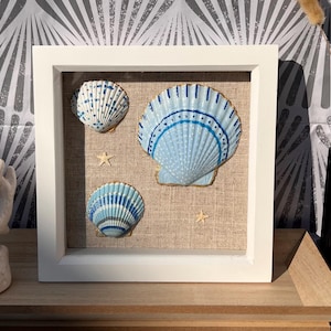 Puede incluir: Un marco cuadrado blanco tipo caja con tres conchas marinas y dos pequeñas estrellas de mar montadas sobre un fondo de lino marrón claro. Las conchas están pintadas en tonos azules y blancos con bordes dorados, dispuestas decorativamente.