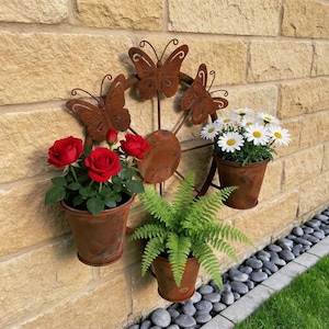 Puede incluir: Un macetero de pared de metal oxidado con diseño de mariposa, con tres plantas en maceta. Las plantas incluyen rosas rojas, margaritas blancas y helechos verdes. El macetero está montado en una pared de ladrillo beige.