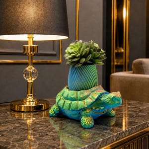 May include: A decorative turtle-shaped planter in shades of blue and green, holding a green succulent plant. The planter sits on a dark marble surface, with a gold lamp in the background.