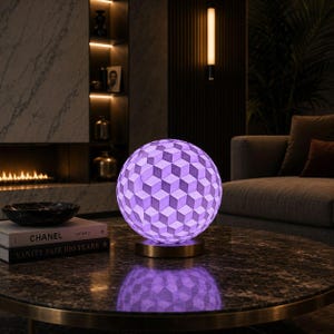 May include: A spherical table lamp with a geometric cube pattern in shades of purple and white. The lamp sits on a gold-colored base and illuminates the surrounding area. The lamp is on a dark marble table.