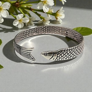 Könnte beinhalten: Ein silberfarbenes Armreif-Armband in Form eines Fisches. Das Armband hat ein Fischschwanzende und einen Fischkopf am anderen Ende, mit detaillierten Schuppen- und Flossenmustern. Das offene Design ermöglicht ein einfaches Tragen.