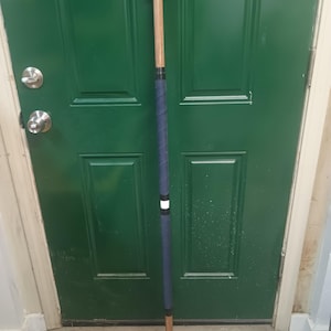 May include: A long wooden staff featuring a natural light brown finish, topped with a blue cap. The centre section is wrapped in a dark blue textured grip, accented by a small white band near the top of the handle, and finished with a blue base.