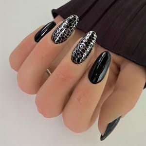Peut inclure: Ongles en forme d'amande avec une finition noire brillante sur les ongles extérieurs et un motif métallique argenté et noir rappelant une peau de serpent sur les deux ongles du milieu. Les ongles sont longs, manucurés et très brillants.
