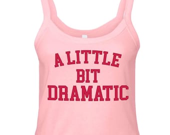 May include: A light pink sleeveless tank top with a scoop neckline and thick shoulder straps. The front of the garment features the text A LITTLE BIT DRAMATIC printed in a bold, dark red, collegiate-style font across the center of the chest.