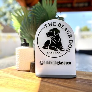 May include: A white, arched acrylic sign featuring a black circular logo of a dog in profile. The text reads THE BLACK DOG LASER CO. and @blackdoglaserco. The sign is mounted on a thin, dark base, presenting a clean, professional display piece.