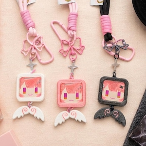 May include: Three lanyards with keychains featuring square frames containing a stylized character face with pink hair and yellow eyes. Each keychain has a bow-shaped clasp, a small star charm, and a pair of wings at the bottom. The lanyards are pink and black.