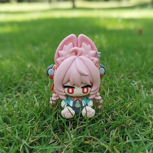 May include: A small, pink figurine of a character with large ears and red eyes, sitting on green grass. The figure has a detailed outfit with teal and black accents. The overall design suggests a collectible or decorative item.