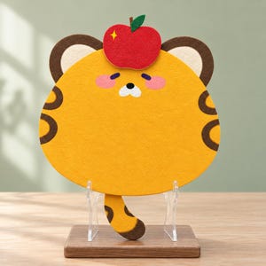 May include: A yellow felt tiger-shaped mat featuring brown ear details, dark brown side stripes, pink cheeks, and a red apple accent on top. The mat is displayed on a clear acrylic stand with a rectangular wooden base.