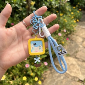 May include: A light blue lanyard with a white wrapped section and a white plastic clip. Attached is a yellow square charm featuring a cartoon face, a small white flower charm in a brown pot, and a blue butterfly-shaped metal connector.
