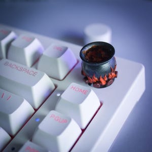 Peut inclure: Un petit capuchon de touche en forme de chaudron noir avec des flammes orange à la base, rempli d'une substance brun foncé, est posé sur un clavier blanc. Accessoire décoratif pour claviers mécaniques.