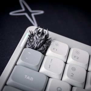 May include: A metallic silver artisan keycap featuring a detailed, spiky, armored helmet design, positioned on a white computer keyboard next to the Tab key. The keycap has a dark, weathered finish with intricate ridges and sharp, pointed elements.