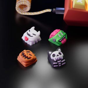 Op de afbeelding: Vier toetsenbordknoppen met Halloween-thema: een witte geest met een rood hart, een groene zombie met een roze hersenen en tong, een oranje pompoen en een zwarte kat met een schedelontwerp. De toetsen zitten op een zwart oppervlak.