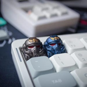 Può includere: Due tasti personalizzati a forma di elmi da soldato futuristici, uno in antracite metallizzato e uno in blu metallizzato, entrambi con emblemi di teschi alati dorati sulla fronte e lenti della visiera rosse, installati su una tastiera bianca.