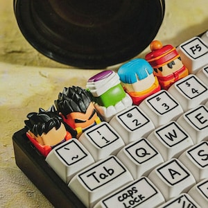 May include: A mechanical keyboard featuring custom artisan keycaps shaped like anime character heads. The white keys have black lettering including Tab, Caps Lock, and numbers. The character keycaps are colorful, featuring black, green, blue, and red hair.
