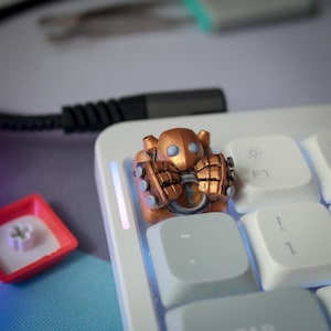 May include: A custom artisan mechanical keyboard keycap featuring a metallic copper-colored robot character with two light blue circular eyes and large mechanical hands, positioned on a white keyboard next to a red keycap base.