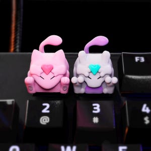 Puede incluir: Dos teclas con forma de gato en rosa y lavanda, con detalles de corazón. La tecla rosa tiene un corazón rosa, y la tecla lavanda tiene un corazón turquesa. Ambas tienen una cola curvada y están en un teclado negro.
