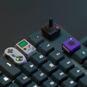 May include: A black mechanical keyboard featuring custom artisan keycaps shaped like retro gaming consoles and controllers, including a gray handheld device, a gray controller, a black joystick, and a purple cube-shaped console.