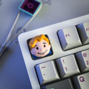 May include: A custom keyboard keycap featuring a cartoon character's face with yellow hair and a smiling expression. The keycap is installed on a white keyboard, next to other keys with purple lettering. A keycap puller is in the background.