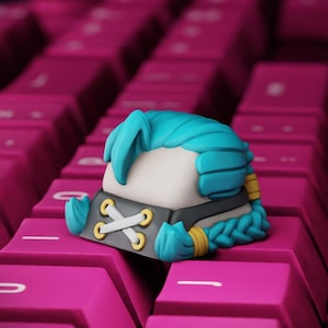 May include: A custom mechanical keyboard keycap featuring a sculpted character head with bright blue hair, a white face, and a black collar detail with gold eyelets and white crisscross lacing. The hair is styled in a braid with gold bands.