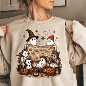 May include: A beige crewneck sweatshirt featuring a graphic illustration of ghosts, vampires, and a creature holding a map labeled Treasure Map of Ghostly Cove. The design includes pumpkins, cameras, and stars in shades of brown, orange, and black.
