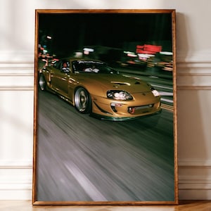 May include: A framed wall art print featuring a gold Toyota Supra sports car driving on a road at night. The car is captured in motion with a blurred background, showing its headlights, front bumper, and a white decal on the windshield.