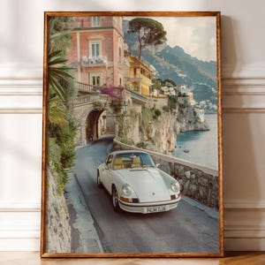 May include: A framed wall art print depicting a white vintage sports car driving on a coastal road. The scene features a stone archway, pastel-colored Mediterranean buildings, lush greenery, and a cliffside overlooking the sea. The car license plate reads PALMS CLUB.