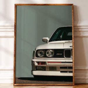 Pode incluir: Uma impressão de arte de parede emoldurada apresentando a vista frontal de um carro esportivo BMW E30 M3 branco. O veículo exibe seus faróis redondos duplos característicos, uma grade preta com emblema M3 e um para-choque retangular com luzes.