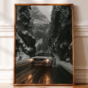 May include: A framed wall art print featuring a white Audi Quattro rally car driving on a wet, dark road through a snowy mountain pass. The car has illuminated yellow headlights and is surrounded by tall, dark pine trees and rocky cliffs.