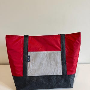 May include: A red and gray tote bag with black accents. The bag features a red top, a gray center pocket, and a black base. Black straps extend from the top to the base. The bag has a rectangular shape and is made of a durable material.