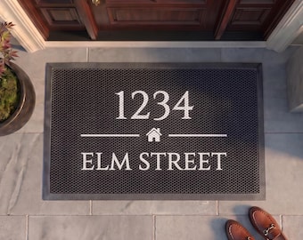 May include: A rectangular dark gray doormat with a textured mesh surface. The mat features white printed text that reads 1234, a small house icon, and the words ELM STREET centered horizontally. The design is clean, functional, and durable.