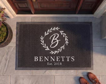 May include: A rectangular black doormat with a textured surface featuring a white circular laurel wreath design surrounding the letter B. Below the wreath, the text reads BENNETTS and Est. 2018 in a clean, white, serif font style.
