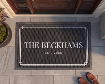 May include: A rectangular black doormat with a textured surface and a white double-line border. The center features the white text THE BECKHAMS and EST. 2026 in a serif font. The mat is positioned on a light gray tiled floor in front of a door.