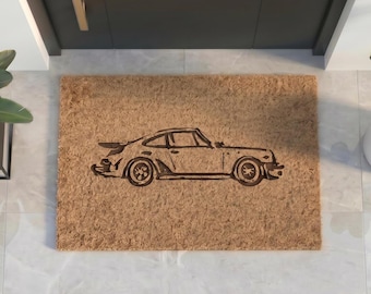 May include: A rectangular tan coir doormat featuring a dark brown, stylized line art illustration of a classic sports car in profile. The car design includes a rear spoiler, circular wheels, and a low-slung body shape centered on the mat.