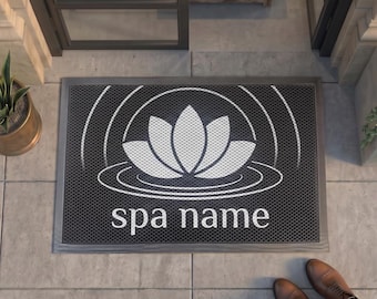 May include: A rectangular black doormat featuring a white graphic of a lotus flower centered above concentric circles. The text "spa name" is printed in white lowercase letters below the lotus design. The mat has a textured surface pattern.