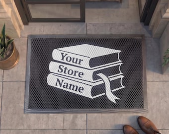 May include: A rectangular black doormat with a textured surface featuring a white graphic of a stack of three books. The text 'Your Store Name' is printed in white on the spines of the books, and a bookmark ribbon hangs from the middle book.