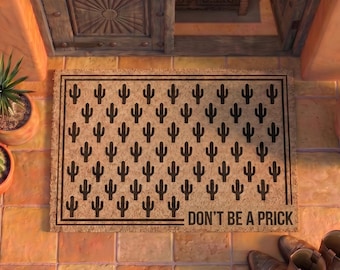 May include: A rectangular tan coir doormat featuring a repeating black cactus pattern and the text DON'T BE A PRICK printed in the bottom right corner. The mat is framed by a thin black border and is positioned on a terracotta tiled floor.