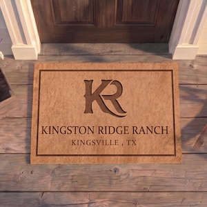 May include: A rectangular brown doormat featuring a dark brown border and a central monogram of the letters KR. The text KINGSTON RIDGE RANCH is printed below the monogram, followed by the location text KINGSVILLE, TX in a dark brown font.