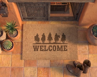 May include: A rectangular tan coir doormat featuring a dark brown graphic of five cowboys on horseback in silhouette above the word WELCOME in a bold, blocky font. The mat is positioned on a terracotta tiled floor near potted plants and boots.