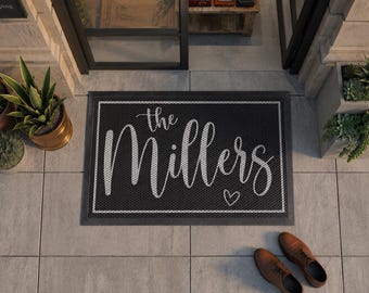 May include: A rectangular black doormat with a white border and white text that reads "the Millers" in a cursive script, accompanied by a small white heart outline. The mat features a textured surface and is positioned on a light gray tiled floor.
