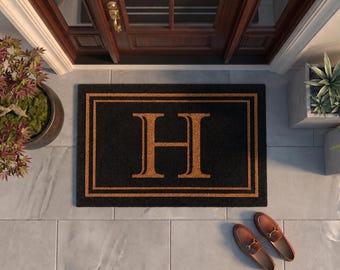 May include: A rectangular black doormat featuring a large, centered tan letter H. The mat is bordered by two thin, tan rectangular lines that frame the letter. The design is consistent with a natural fiber texture, providing a contrast against the dark background of the mat.