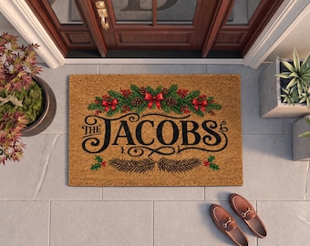 May include: A rectangular tan coir doormat featuring the black text THE JACOBS in a decorative script font. The design includes a festive holiday garland of green pine needles, brown pinecones, red berries, and red bows at the top and bottom.