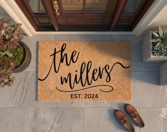 May include: A rectangular tan coir doormat featuring black cursive text that reads the millers and the text EST. 2024 centered below. The mat is positioned on a gray tiled floor in front of a dark brown wooden door with a gold handle and a glass panel.