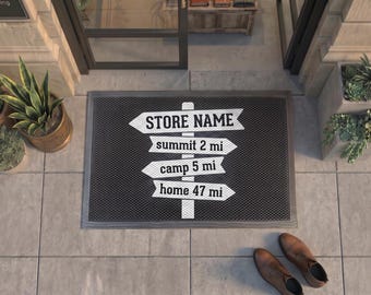 May include: A rectangular black doormat with a white graphic of a vertical signpost featuring four directional arrows. The arrows contain the text STORE NAME, summit 2 mi, camp 5 mi, and home 47 mi in a bold, black, sans-serif font.