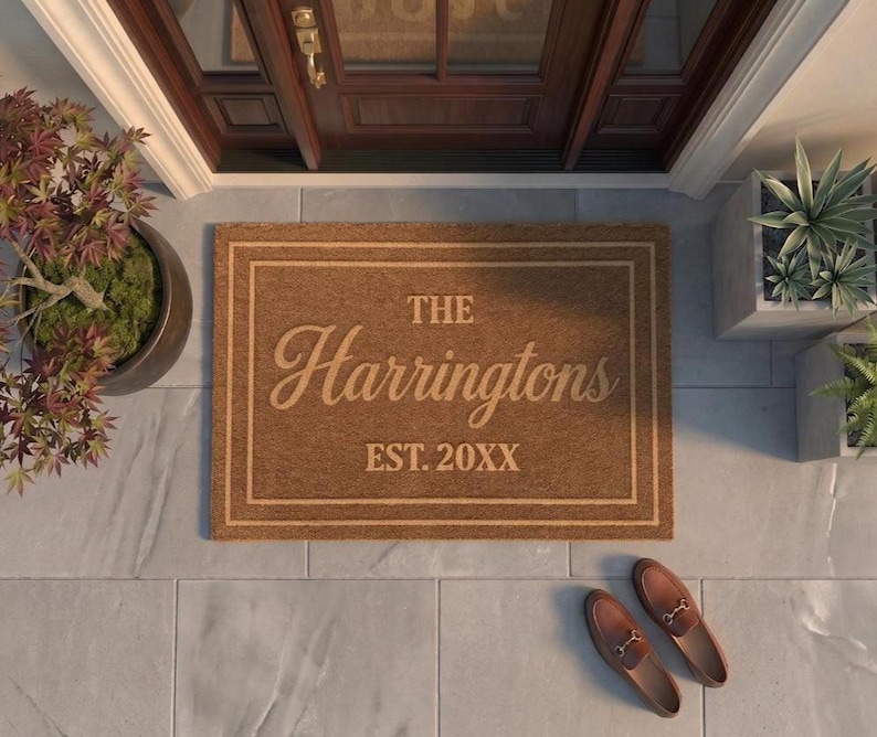 May include: A rectangular brown coir doormat featuring a double-line border and the text THE Harringtons EST. 20XX in a dark brown font. The mat is centered on a light gray tiled floor with a dark wooden door visible in the background.
