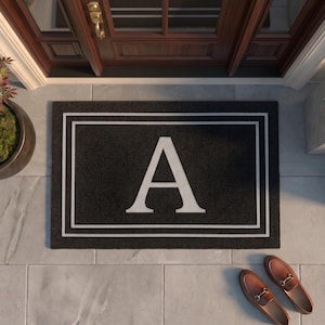 May include: A rectangular black doormat featuring a large white capital letter A centered within a double-lined white rectangular border. The mat is positioned on a light gray tiled surface in front of a dark brown wooden door with glass panels.