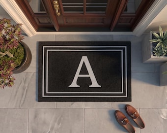 May include: A rectangular black doormat featuring a large white capital letter A centered within a double-lined white rectangular border. The mat is positioned on a light gray tiled surface in front of a dark brown wooden door with glass panels.