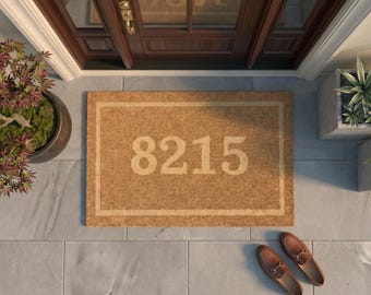 May include: A rectangular tan coir doormat featuring a lighter tan border and the house number 8215 printed in a bold, sans-serif font in the center. The mat has a coarse, natural fiber texture and is positioned on a gray tiled entryway floor.